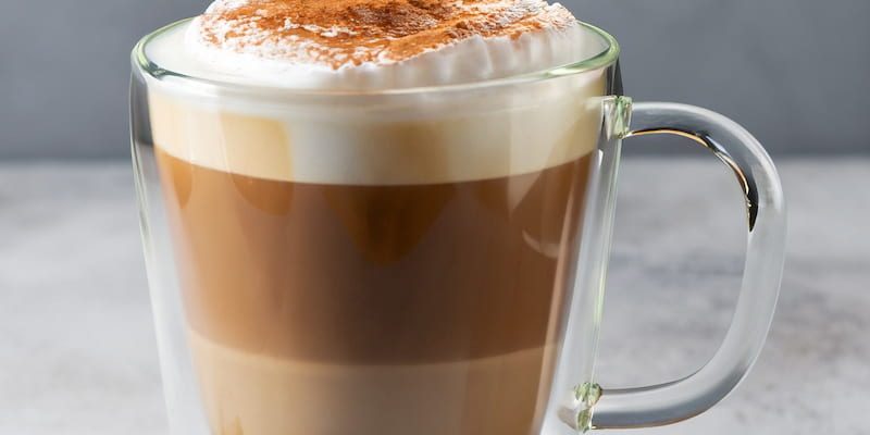 a glass cup containing a latte with milk foam and cinnamon on top, next to cinnamon sticks and coffee beans on a gray surface.
