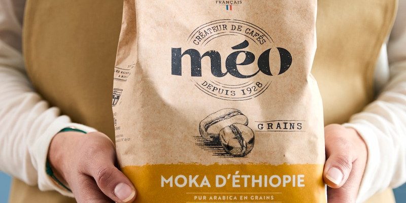 a person in an apron is holding a labeled bag of meo coffee "Ethiopian mocha, pure arabica beans". the bag is decorated with a brown and yellow pattern representing coffee beans.