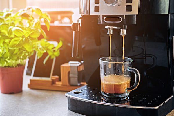 espresso machine brewing coffee in a transparent glass cup, with a basil plant and kitchen utensils in the background.