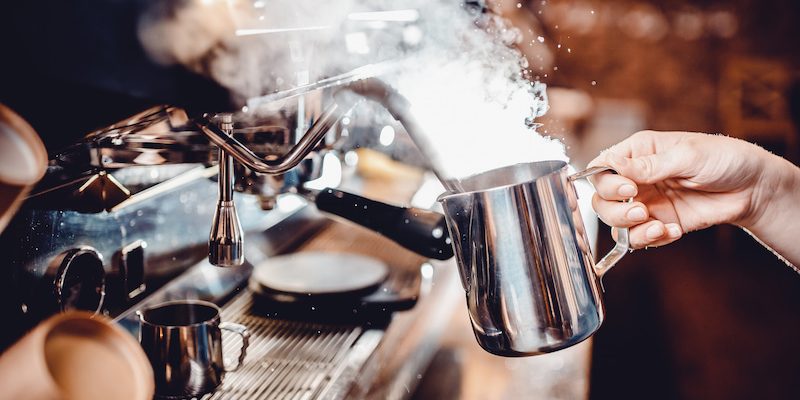 whip milk foam by hand with a pitcher next to an espresso machine, releasing visible steam.